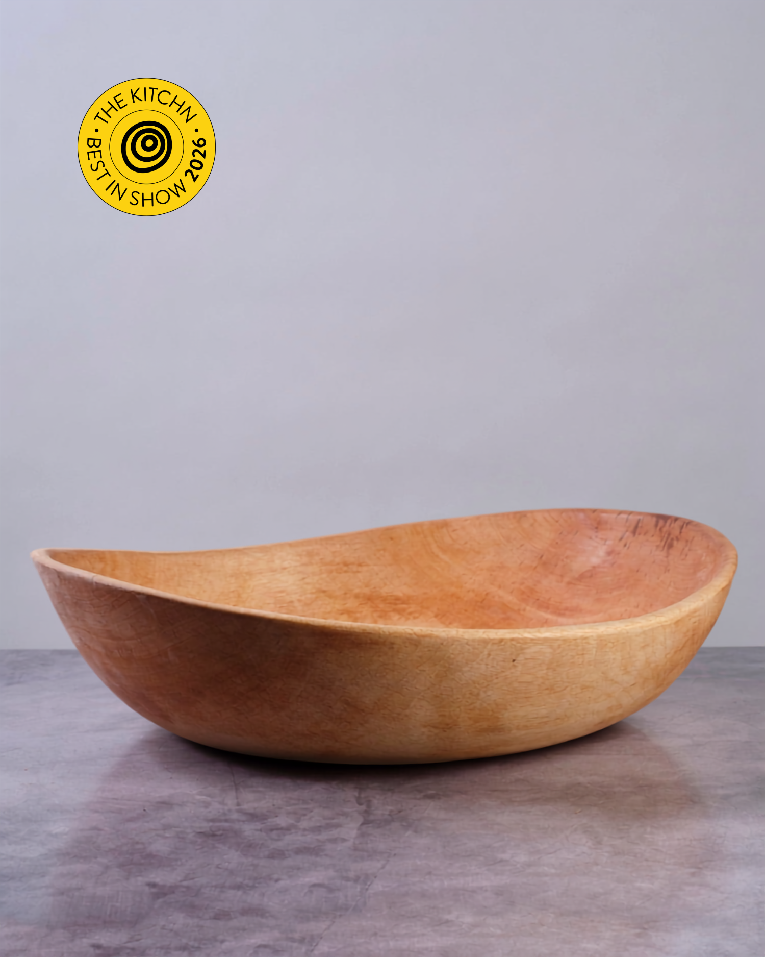A large, organic-shaped wooden bowl with a warm honey-brown finish, set on a gray surface against a neutral gray background. A yellow circular badge in the top left corner reads 'THE KITCHN BEST IN SHOW 2026'.