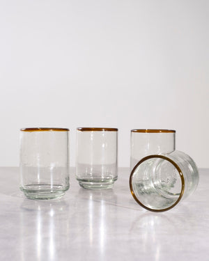 Four clear glass tumblers with a thin amber-brown rim, shown on a light gray reflective surface against a white background. 
