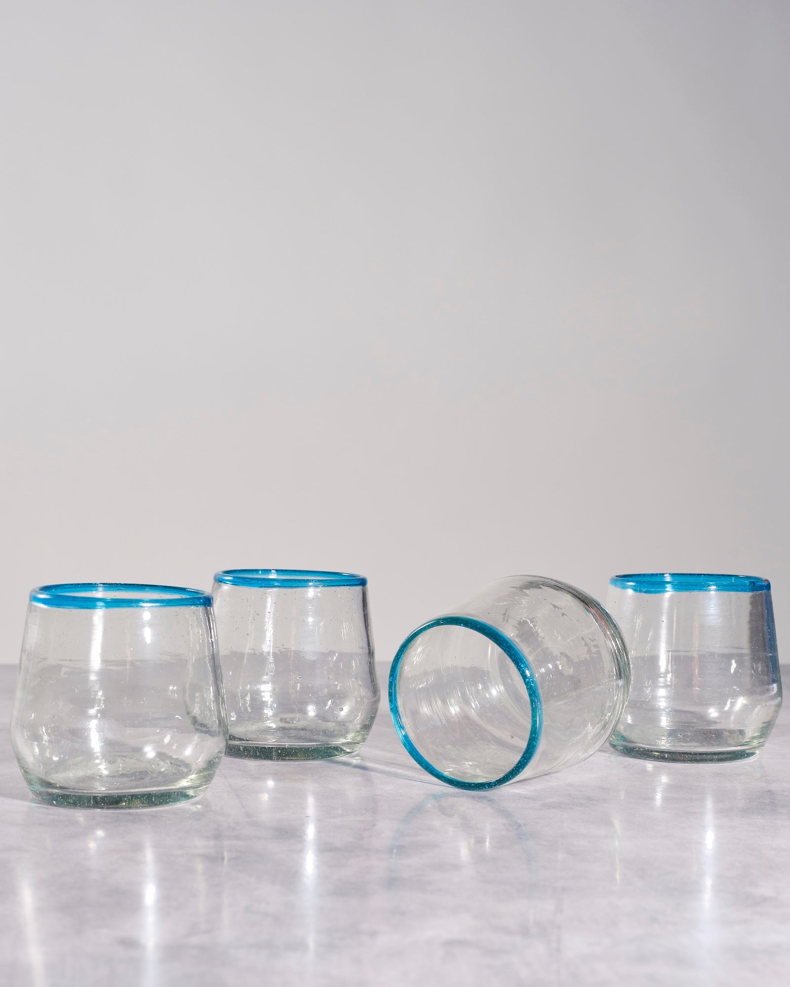 Four clear stemless drinking glasses with bright blue rim detailing, arranged on a light gray marble surface against a neutral light gray background.