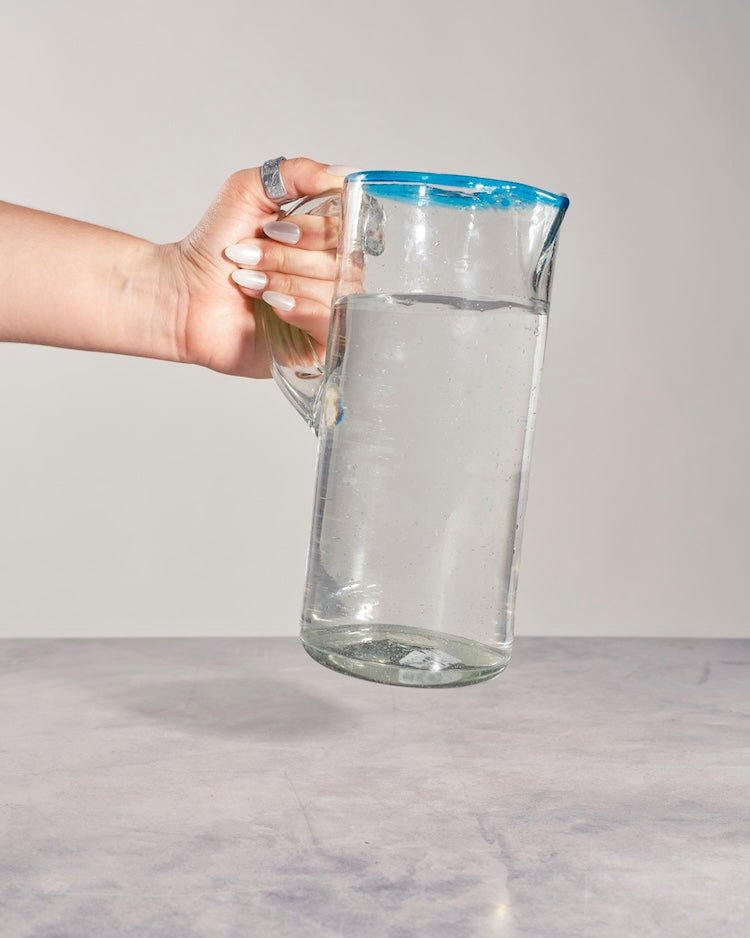 A clear glass pitcher with a blue rim, partially filled with water, held by a person's hand against a light gray background.