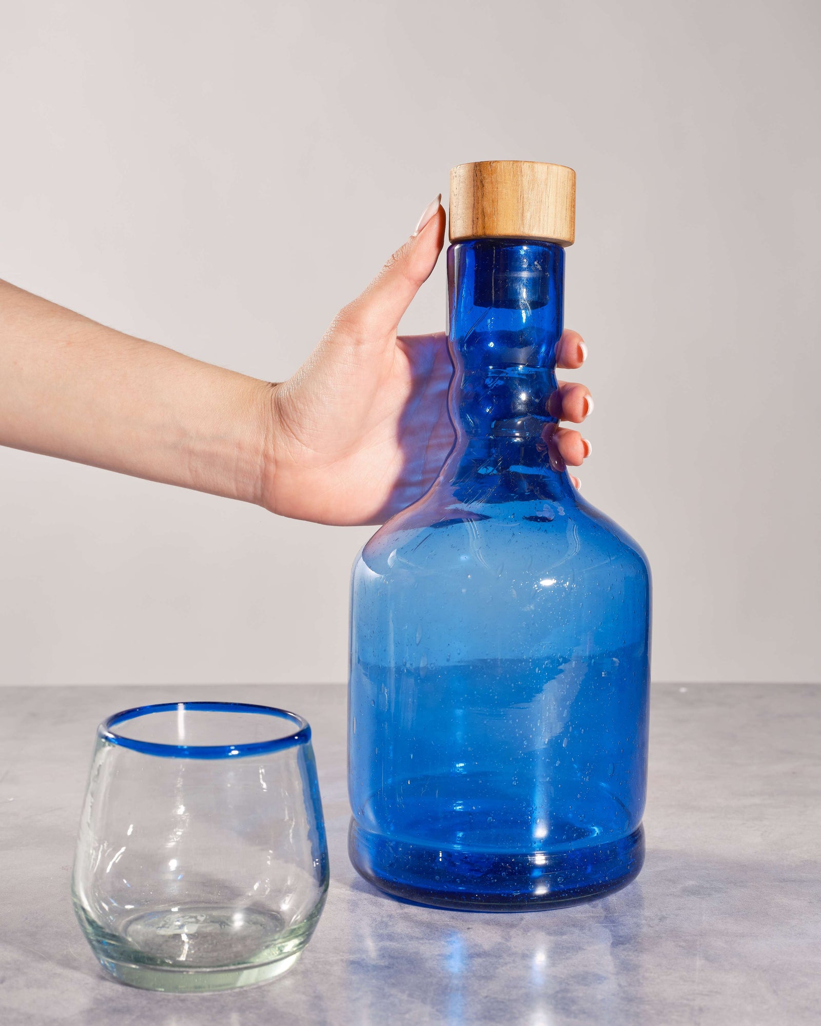TerraKlay's Blue glass bottle with wooden cap held by a hand, next to a clear wine glass on a light gray background.
