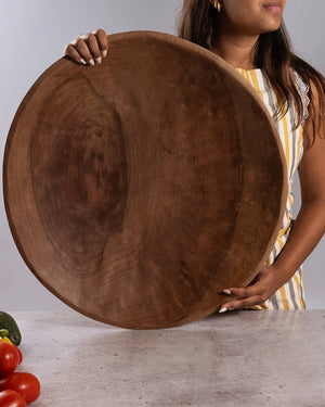 A woman holds a very large round wooden shallow wood bowl or serving platter in front of a light gray studio background, with a few vegetables visible on a tabletop.