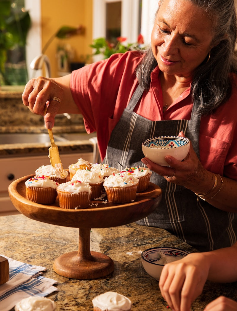 Woman in a kitchen decorating cupcakes on a wooden stand.