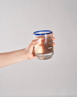 A hand holding a clear glass tumbler with a blue rim, filled with a brown liquid, against a plain light gray background.