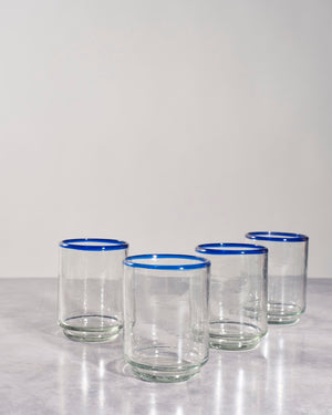 Four clear glass tumblers with a vibrant blue rim, arranged on a light gray marble surface against a neutral light gray background.