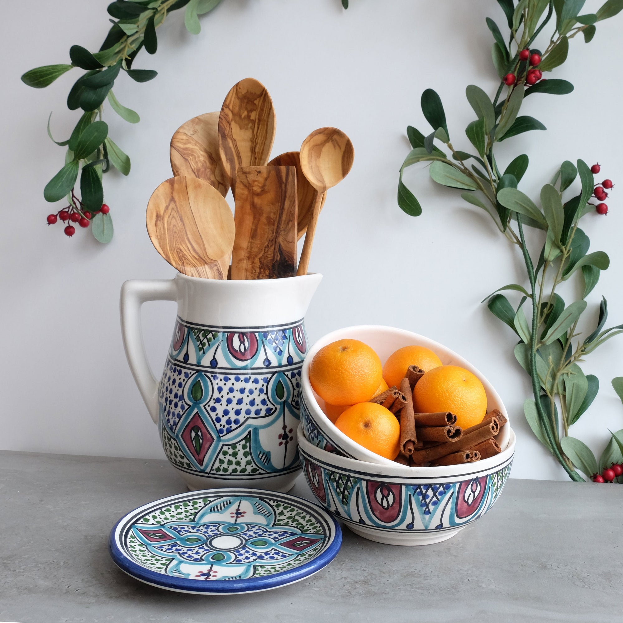 A ceramic pitcher with a colorful hand-painted geometric pattern, accompanied by a wooden spoon, displayed with fruits and spices in the background, suggesting its use for serving.