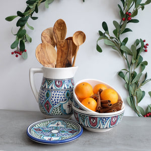 A ceramic pitcher with a colorful hand-painted geometric pattern, accompanied by a wooden spoon, displayed with fruits and spices in the background, suggesting its use for serving.