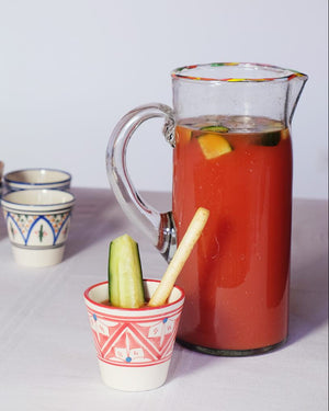 A handblown glass pitcher with a red rim and amber tinted glass, accompanied by small ceramic cups and a slice of lemon, on a white surface.