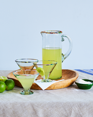 Pitcher and glasses of green liquid on a wooden tray with limes and a bowl of salt.